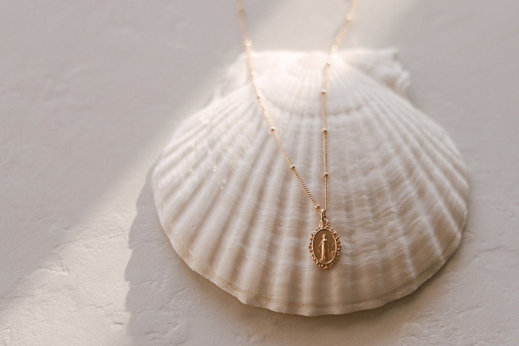 Gold-tone oval medal pendant necklace laid flat on white scallop shell