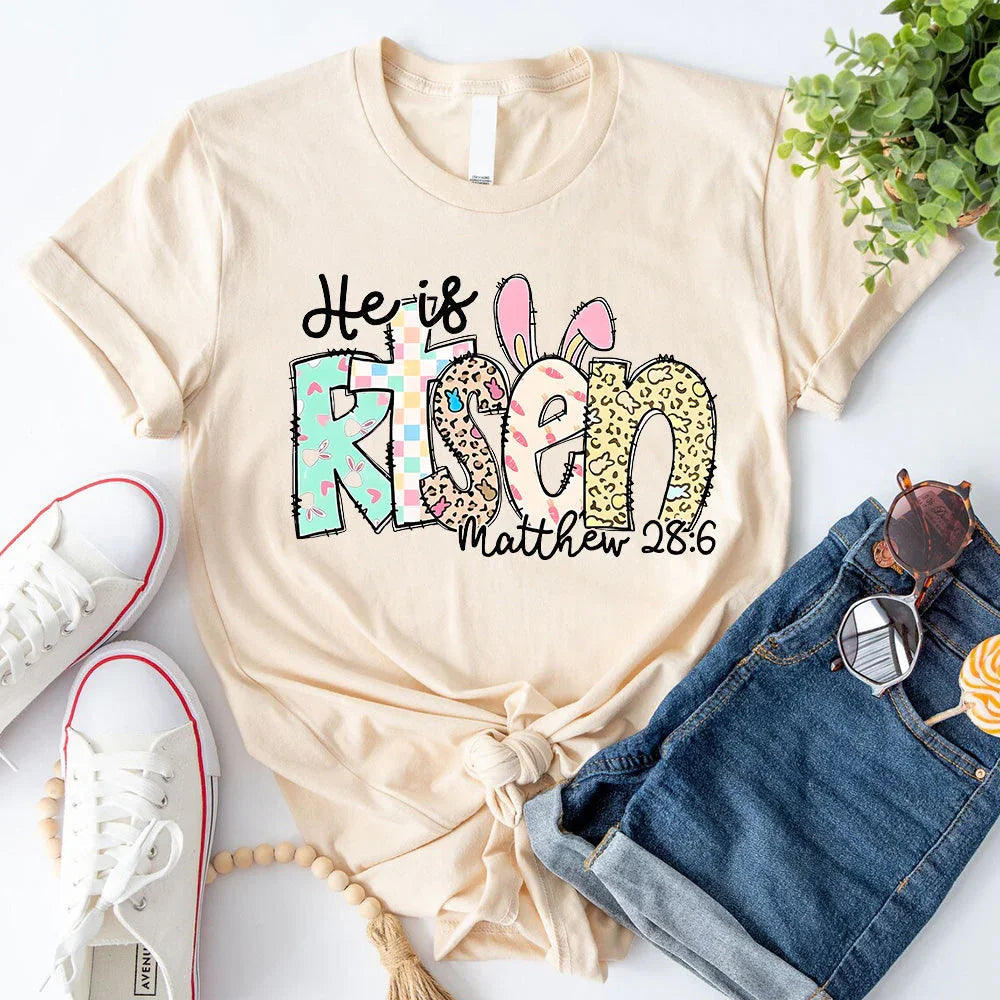 Cream crewneck T-shirt with colorful 'He is Risen' bunny graphic and 'Matthew 28:6' text