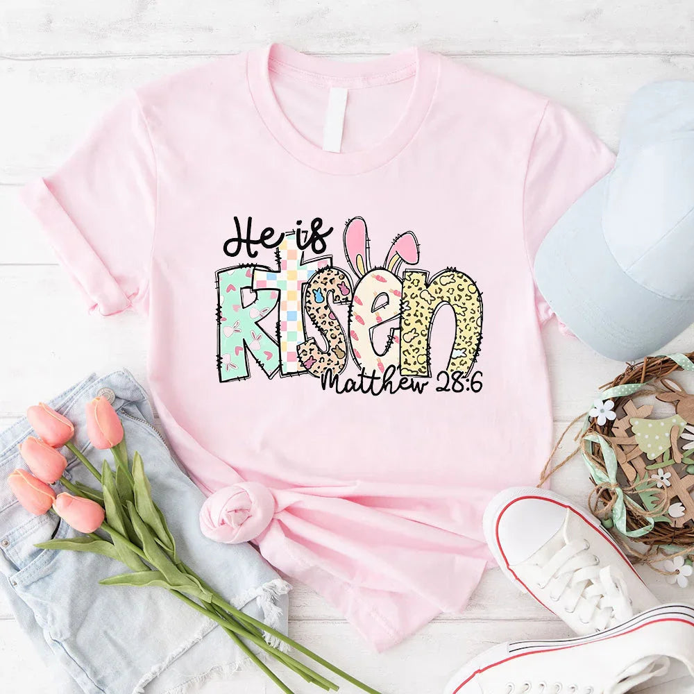 Pink crewneck T-shirt with colorful 'He is Risen' bunny graphic and 'Matthew 28:6' text