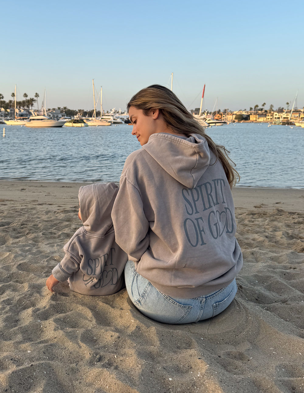 Kids taupe hoodie with hood and “SPIRIT OF GOD” back print, child seated at beach