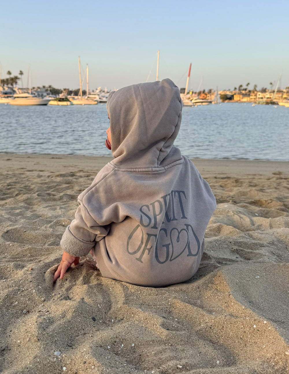 Kids taupe hoodie with hood and “SPIRIT OF GOD” back graphic, child sitting on sand